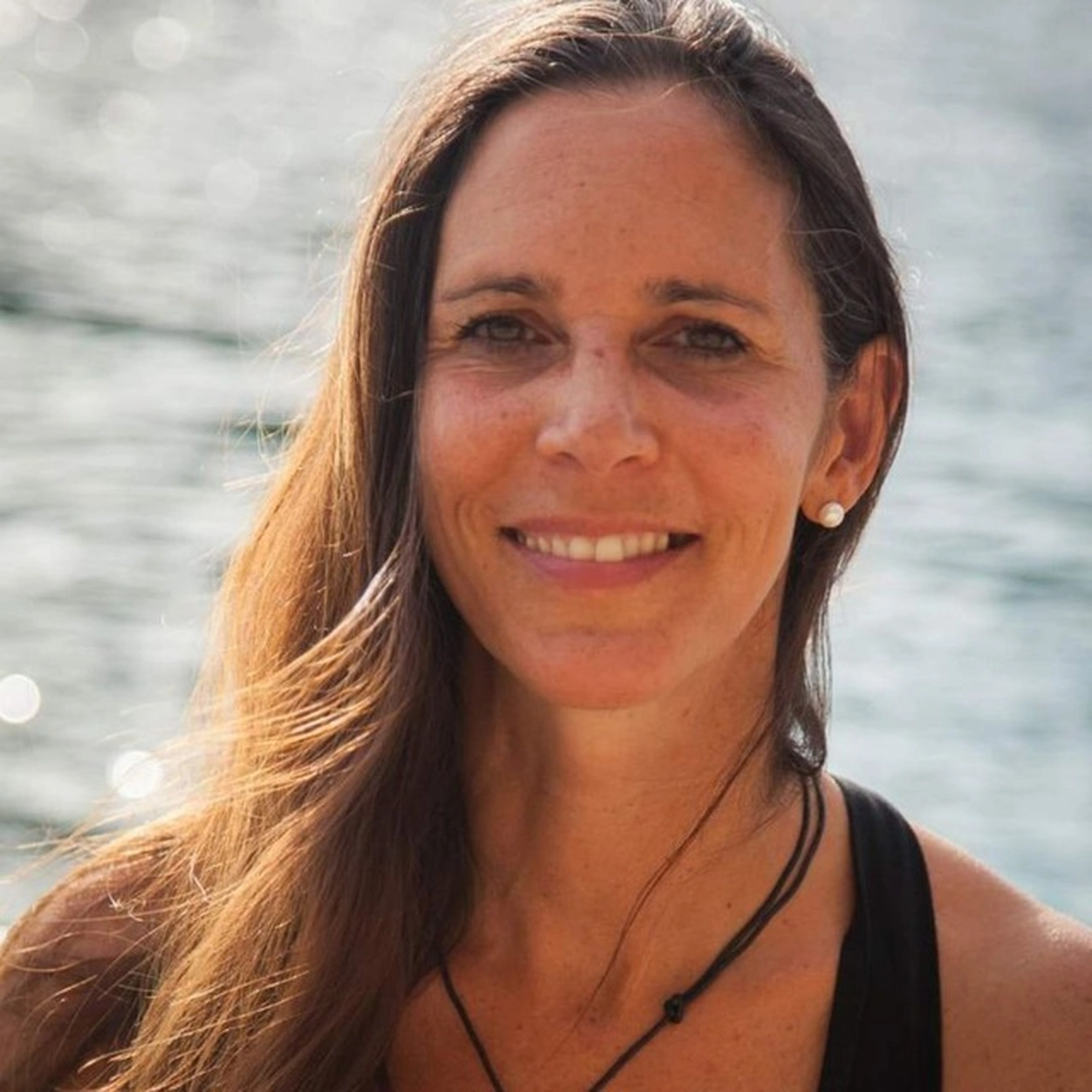 Körperglück -Massage Therapy and Pelvic Floor Training - Wooden tables, chairs, and palm trees on a stone terrace overlooking a black pebble beach and distant sea cliffs under a bright blue sky.