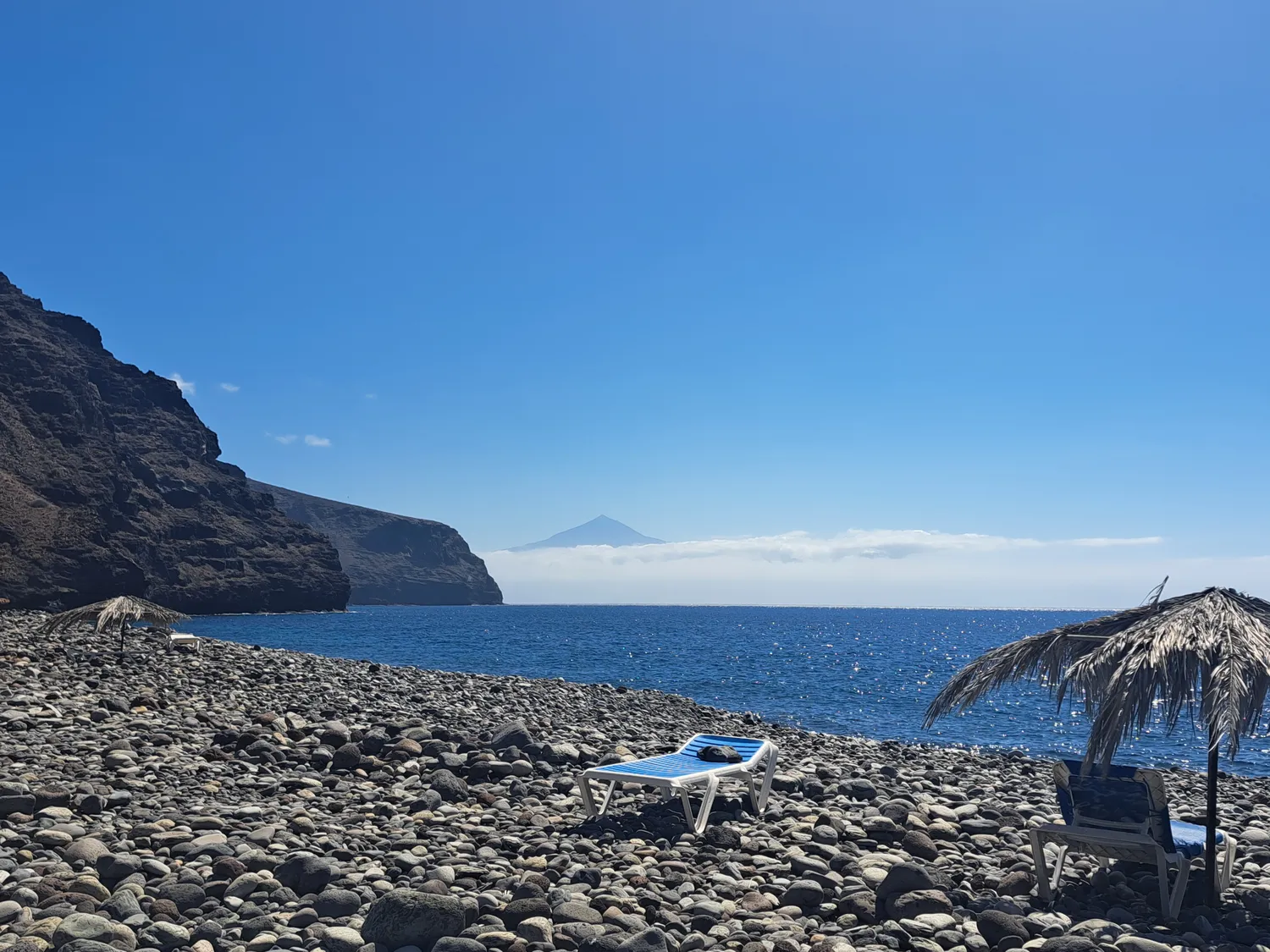 Körperglück - Massagetherapie und Beckenbodentraining - Eine einsame blaue Sonnenliege an einem vulkanischen Kieselstrand mit Blick auf den Ozean. Im Hintergrund erhebt sich ein Berggipfel über einer Wolkenschicht unter klarem, blauem Himmel.