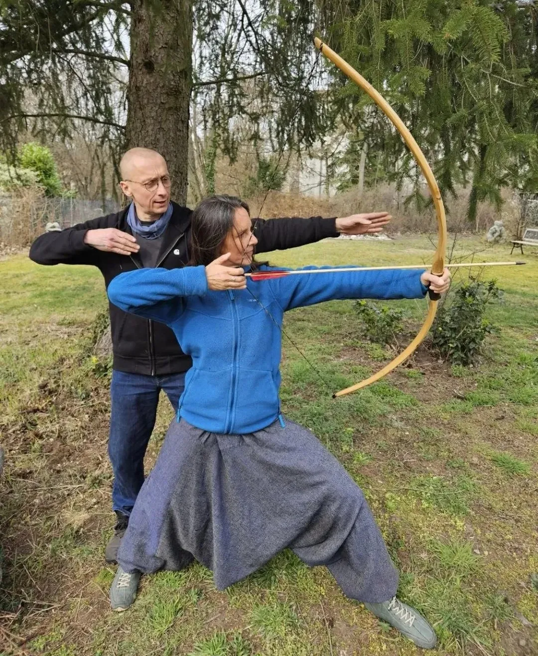 Körperglück - Massagetherapie und Beckenbodentraining - Eine Frau übt im Freien Bogenschießen mit einem Holzbogen, während ein männlicher Trainer hinter ihr steht und ihre Haltung korrigiert.
